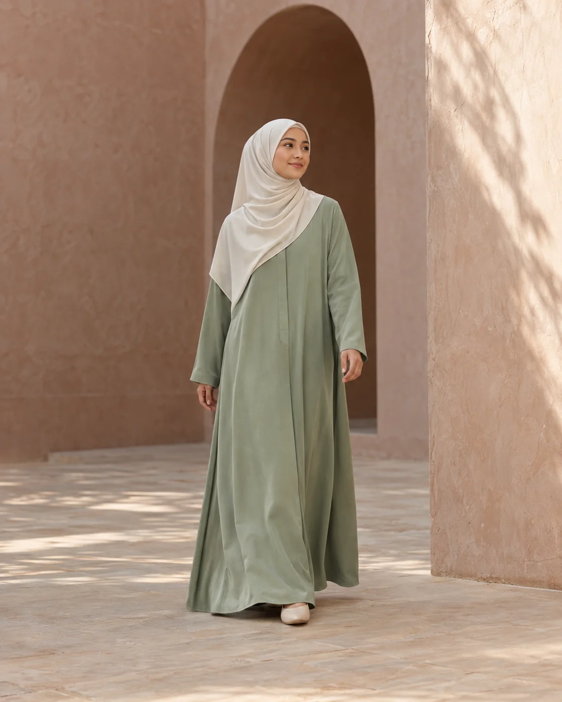 A woman in a flowing sage dress and ivory hijab walking through a sunlit blush courtyard.