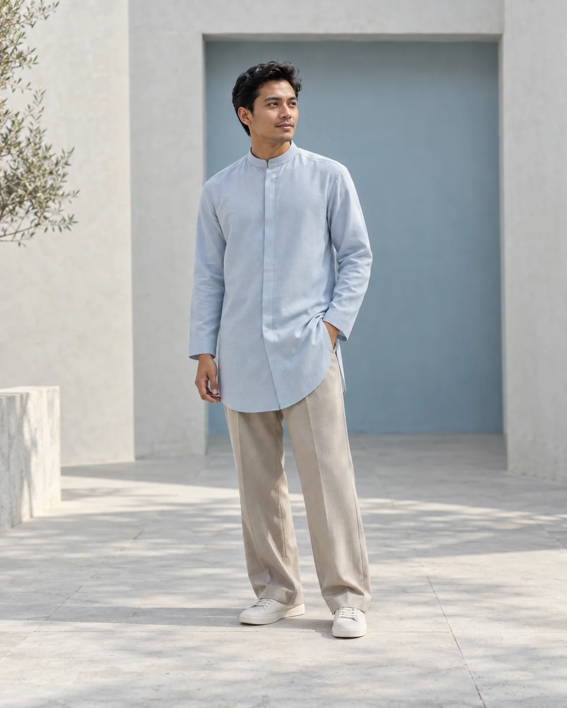 A man in a powder-blue longline tunic and stone trousers beside a cool blue architectural recess.